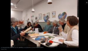 Sieben ältere Frauen sitzen in einem gemütlichen Raum um einen Tisch herum, diskutieren und machen sich Notizen. Karten, Papiere und Kaffeetassen stehen auf dem Tisch. Auf einem Schild an der Wand steht "omas for future".