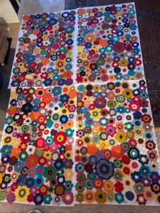 Vier große Tafeln mit bunten, gehäkelten Blumenmotiven sind nebeneinander auf dem Boden ausgelegt und bilden ein lebendiges und kompliziertes Patchwork-Muster, das auf subtile Weise die Themen Natur und Ökozid widerspiegelt. Jedes Feld weist eine Vielzahl von Formen und Farben auf.