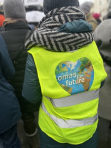 Eine Person, die eine leuchtend gelbe Weste mit einer herzförmigen Weltkugel und der Aufschrift omas for future trägt, steht inmitten einer Menschenmenge, von hinten gesehen. Die Person trägt außerdem eine graue Strickmütze und einen gestreiften Schal.