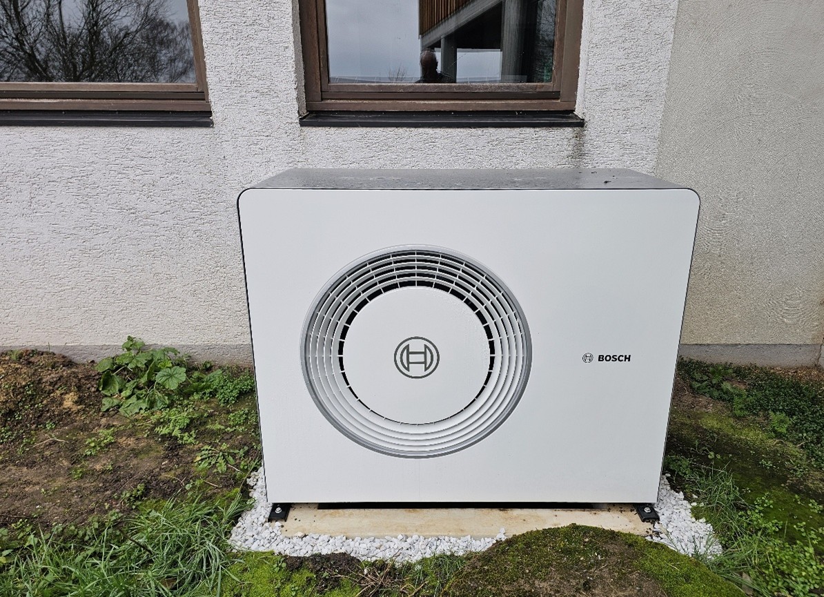 Ein Bosch Wärmepumpe-Außengerät ist neben einer Hauswand auf einer Betonplatte installiert, die von Gras und Erde umgeben ist. Das Gerät ist mit einem runden Lüftungsschlitz und dem Bosch-Logo auf der Vorderseite versehen und liefert erneuerbare Energie für Ihr Haus.