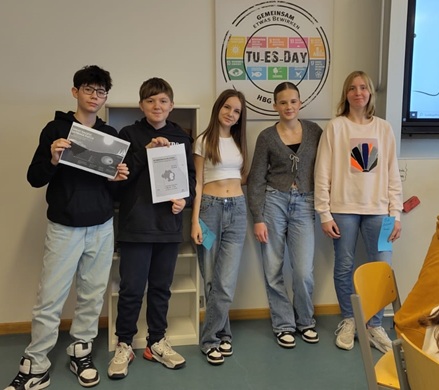 Fünf Jugendliche stehen Seite an Seite in einem Klassenzimmer. Zwei von ihnen halten Zeugnisse in der Hand. Alle sind leger gekleidet, drei von ihnen tragen hellblaue Jeans. An der Wand hinter ihnen ist ein rundes Plakat mit der Aufschrift TUESDAY zu sehen.