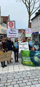 Eine Gruppe von Menschen steht und kniet auf einer Kopfsteinpflasterstraße und hält Schilder zum Klimaschutz, zur Demokratie und zum Umweltschutz in die Höhe. Im Hintergrund sind Gebäude, ein Schild mit einer 20 km/h-Zone und ein blattloser Baum zu sehen.