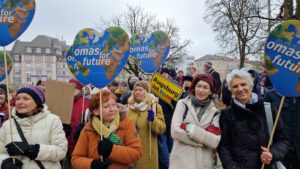 Eine Gruppe älterer Frauen hält bei einem Protest herzförmige Schilder mit der Aufschrift Omas for Future. Sie tragen Winterkleidung und stehen im Freien mit anderen Demonstranten und Bäumen im Hintergrund. Es sind Mitglieder der Regionalgruppe Augsburg
