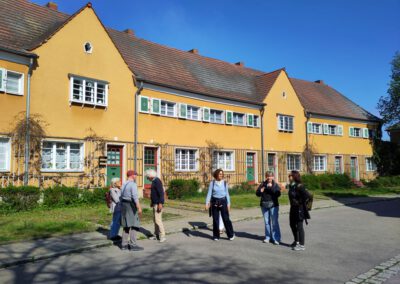 Eine Gruppe von sechs Personen steht und unterhält sich auf einer sonnigen Straße vor einer Reihe gelber, zweistöckiger Häuser mit roten Ziegeldächern und grünen Türen. Der Himmel ist klar und blau, und die Szene wirkt ruhig und freundlich.