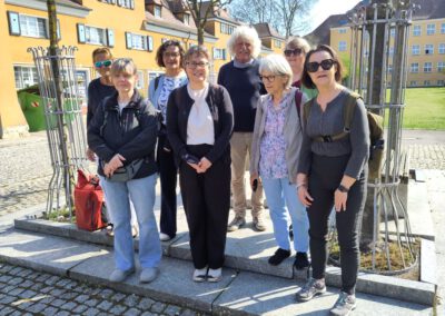 Eine Gruppe von acht Erwachsenen, einige mit Sonnenbrillen, steht an einem sonnigen Tag im Freien vor gelben Gebäuden mit roten Dächern. Sie lächeln in die Kamera, umgeben von Bäumen und grünem Gras.