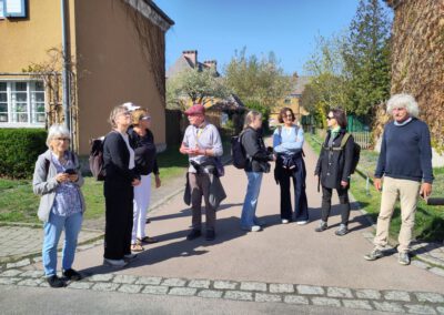 Eine Gruppe von neun Erwachsenen steht plaudernd auf einer sonnigen Straße, die von bunten Häusern und Bäumen gesäumt ist. Einige von ihnen halten Taschen in der Hand und tragen Sonnenbrillen oder Hüte, wobei die Schatten auf helles Tageslicht hinweisen.