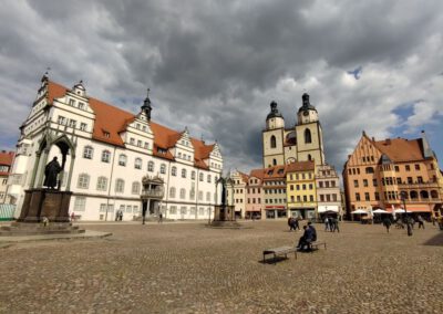 Ein großer kopfsteingepflasterter Platz in einer europäischen Stadt, umgeben von historischen Gebäuden mit roten Dächern und hohen Kirchtürmen; Menschen gehen spazieren und sitzen auf Bänken unter einem bewölkten Himmel.