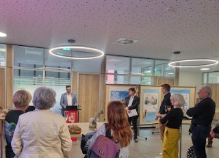Ein Mann spricht auf einem Podium zu einem kleinen Publikum in einem modernen Raum mit Holzwänden; in der Nähe sind Kunstwerke auf Staffeleien ausgestellt, und kreisförmige Deckenleuchten beleuchten die Szene.