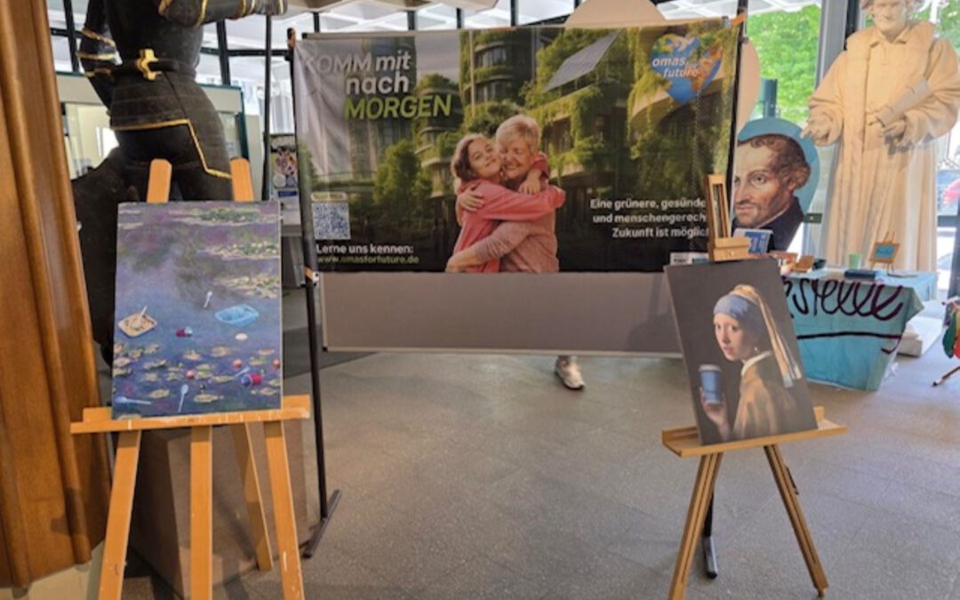 In einem Innenraum sind zwei Gemälde auf Staffeleien ausgestellt. Dahinter befindet sich ein Transparent mit zwei Kindern und einem deutschen Text. Im Hintergrund sind Schaufensterpuppen in historischen Kostümen zu sehen. Von außen fällt etwas natürliches Licht ein.