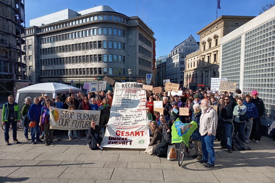 Eine große Gruppe von Menschen versammelt sich an einem sonnigen Tag im Freien mit verschiedenen Protestschildern, darunter Stop burning our future, vor städtischen Gebäuden. Die Stimmung ist ausgelassen und die Gruppe ist bunt gemischt.