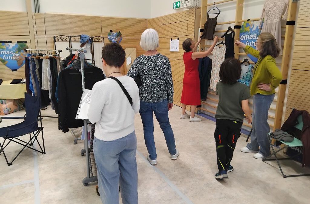 In einem Raum mit hellen Wänden und Holzpaneelen stöbern fünf Personen in Kleiderständern und Wandauslagen. Einige sehen sich Kleider an, während andere die Kleidung auf Kleiderbügeln begutachten. Im Hintergrund sind Schilder und Stühle zu sehen.