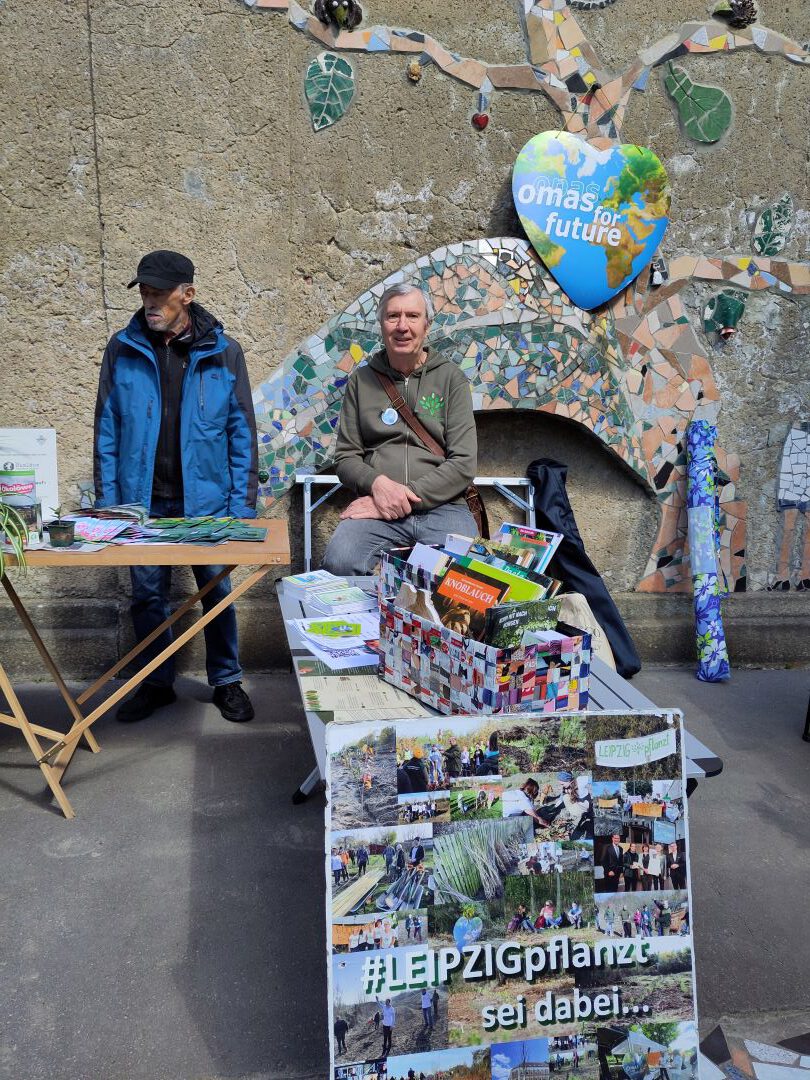 Zwei ältere Männer stehen und sitzen hinter einem Tisch mit Broschüren und Pflanzen bei einer Veranstaltung im Freien. Eine dekorierte Wand mit einem herzförmigen "omas for future"-Schild und eine Collagetafel mit der Aufschrift #LEIPZIGpflanze sind im Hintergrund zu sehen.