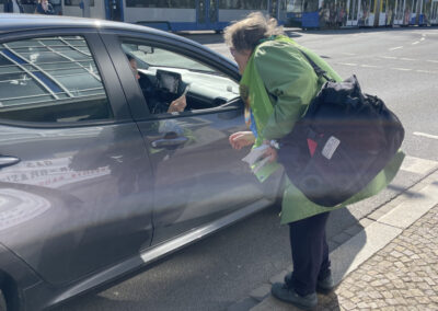 Eine Person in grüner Jacke und mit einer großen Tasche lehnt sich an das Fenster eines geparkten grauen Autos und spricht mit dem Fahrer in einer sonnigen Straße, in der im Hintergrund Straßenbahnen zu sehen sind.