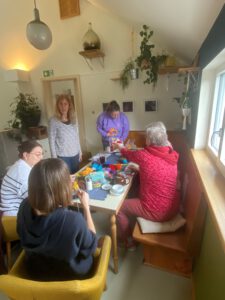 Fünf Frauen sitzen und stehen um einen mit Bastelmaterial, Snacks und Getränken gedeckten Esstisch in einem gemütlichen, mit Pflanzen gefüllten Raum, in den natürliches Licht durch das Fenster fällt.