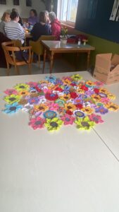 Auf einem großen Tisch sind bunte, gehäkelte Blumenmotive in einem Muster angeordnet. Im Hintergrund sitzen mehrere Personen an einem Tisch in der Nähe eines Fensters und unterhalten sich. Auf der rechten Seite des Tisches steht eine Pappschachtel.