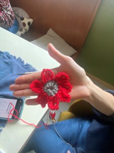 Eine Person hält eine gehäkelte Blume mit roten Blütenblättern und einer grauen Mitte. Die Blume wird gerade gehäkelt, wobei eine Häkelnadel und rotes Garn in der Nähe auf einem Tisch zu sehen sind.