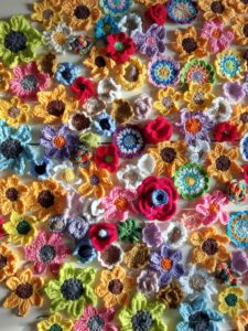Eine farbenfrohe Auswahl an gehäkelten Blumen in verschiedenen Formen und Größen, die dicht nebeneinander auf einer flachen Oberfläche angeordnet sind und leuchtende Gelb-, Rot-, Blau-, Grüntöne und andere kräftige Farben zeigen.