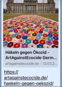 Ein großes Display aus bunten gehäkelten Blumen bedeckt den Boden vor einem historischen Gebäude und wirbt für die Initiative Häkeln gegen Ökozid von Art Against Ecocide Germany.