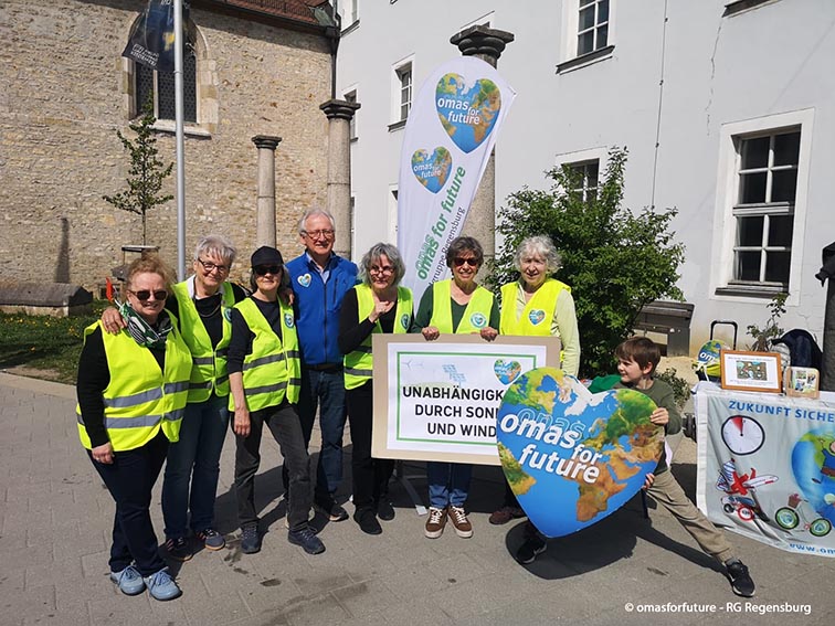 Eine Gruppe von Menschen mit gelben Westen steht im Freien, lächelt und hält Schilder, die für Omas for Future und erneuerbare Energien werben. Ein Kind steht in der Nähe neben einem Globus und einem Schautisch mit Informationsmaterial.