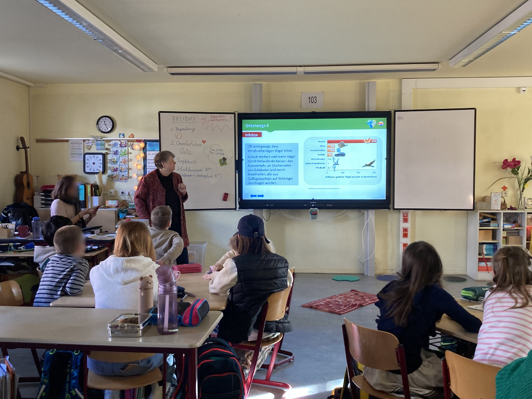 Ein Klassenzimmer mit Schülern, die an Tischen sitzen und einem Lehrer gegenübersitzen, der eine Unterrichtsstunde auf einem großen digitalen Bildschirm präsentiert.