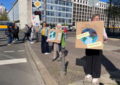 Drei Frauen stehen auf einer Stadtstraße und halten Protestschilder mit Abbildungen der Erde und Klimabotschaften. Im Hintergrund sind andere Menschen und Busse an einem sonnigen Tag zu sehen.