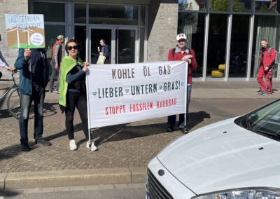 Drei Personen stehen auf einem Bürgersteig und halten ein Transparent mit der Aufschrift "Kohle Öl Gas lieber unterm Gras! Stoppt fossilen Raubbau" bei einer Protestaktion. Ein weiteres Schild in deutscher Sprache wird in der Nähe gehalten. Im Vordergrund ist ein weißes Auto zu sehen.