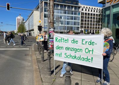 Eine Gruppe von Menschen protestiert auf einer Straße in der Stadt mit einem großen weißen Transparent, auf dem steht: Rettet die Erde - den einzigen Ort mit Schokolade! (Rettet die Erde - der einzige Ort mit Schokolade!). Im Hintergrund sind weitere Schilder zu sehen.