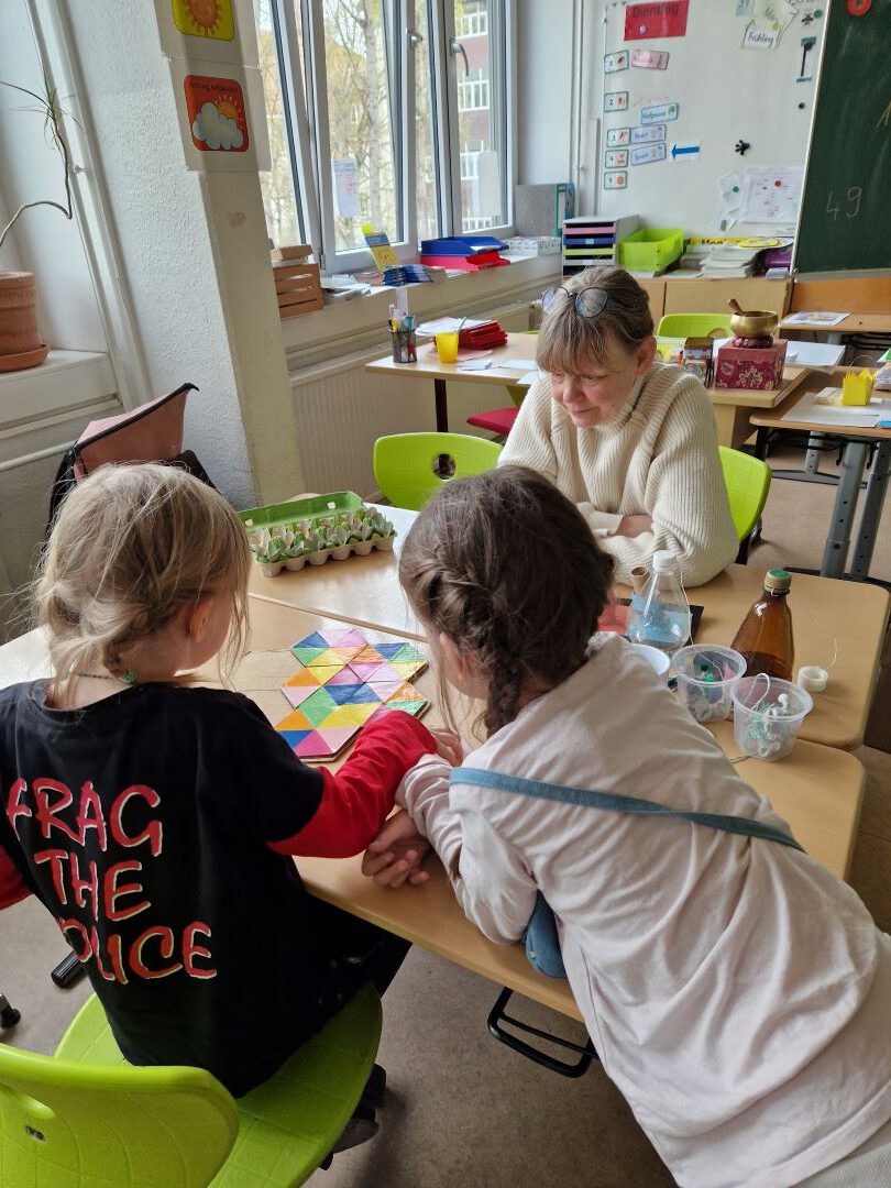 Zwei Kinder arbeiten an einem Klassenzimmertisch an einem bunten Puzzle, während ein Erwachsener lächelnd zusieht. Das Klassenzimmer ist hell mit großen Fenstern, grünen Stühlen und verschiedenen Gegenständen und Dekorationen an den Wänden und Tischen.