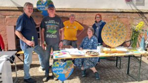 Fünf Personen an einem Informationsstand von Omas for Future im Freien. Drei stehen hinter dem Tisch, zwei sitzen, mit Flugblättern, Pflanzen und einem Spinnrad im Blick. Auf dem Tisch steht ein herzförmiges Erdzeichen.