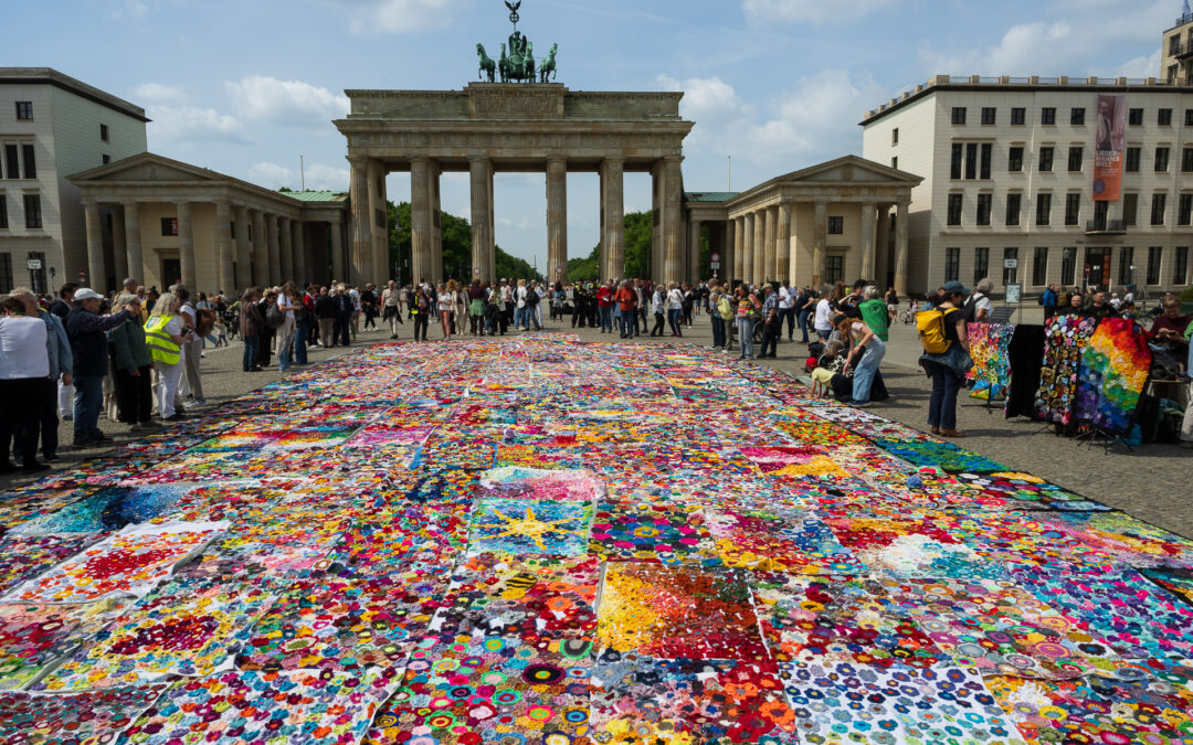 Eine große, bunte Patchworkdecke bedeckt den Boden vor dem Brandenburger Tor in Berlin, umgeben von Menschenmengen unter einem teilweise bewölkten Himmel.