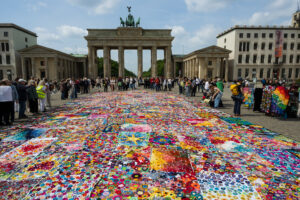 Eine große, bunte Patchworkdecke bedeckt den Boden vor dem Brandenburger Tor in Berlin, umgeben von Menschenmengen unter einem teilweise bewölkten Himmel.