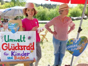 Zwei ältere Frauen stehen an einem Veranstaltungsstand im Freien unter Regenschirmen. Die eine hält ein Schild mit der Aufschrift Umwelt Glücksrad für Kinder und Basteln, daneben stehen herzförmige Omas for Future-Schilder. Es ist ein sonniger Tag mit viel Grün im Hintergrund.