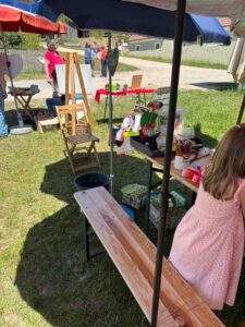 Kunsthandwerksbereich im Freien mit Tischen mit Material unter Sonnenschirmen. Ein Kind in einem rosafarbenen Kleid sitzt auf einer Bank, auf der Kunstmaterialien verstreut sind. Staffelei, Menschen und weitere Tische sind im sonnigen Hintergrund zu sehen.