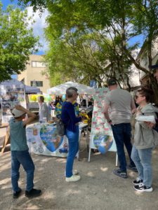 Menschen stehen und unterhalten sich an einem Stand von Omas for Future im Freien, mit bunten Schildern und Plakaten unter belaubten Bäumen. Ein Kind steht in der Nähe, und im Hintergrund sind an einem sonnigen Tag mehrere Stände aufgebaut.