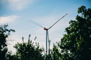Eine hohe Windkraftanlage mit drei Flügeln steht hinter üppig grünen Bäumen unter einem blauen Himmel mit leichten Wolken in Uetersen und zeigt das Engagement von Green Planet Energy für Stromwechsel und Nachhaltigkeit.