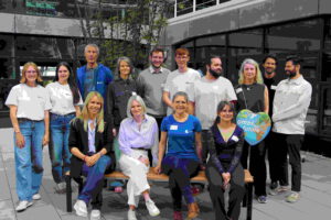 Eine Gruppe von vierzehn Personen, einige stehend und einige auf einer Bank sitzend, posiert und lächelt für ein Foto im Freien in einem modernen baumentscheid münchener Innenhof. Eine Person hält ein buntes herzförmiges Schild in der Hand.