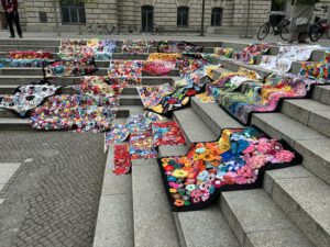 Bunte gehäkelte Decken und Stücke mit Blumenmustern sind auf breiten Steinstufen im Freien ausgestellt und bilden einen lebhaften Kontrast zu den grauen Stufen und dem Bürgersteig. Im Hintergrund sind Fahrräder und ein Gebäude zu sehen.