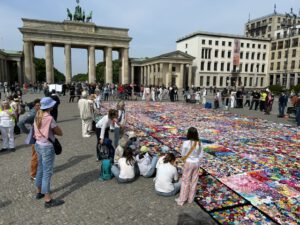 Eine große Gruppe von Menschen versammelt sich vor dem Brandenburger Tor in Berlin und betrachtet und gestaltet eine bunte Patchwork-Kunstinstallation, die auf dem Boden des offenen Platzes ausgelegt ist.