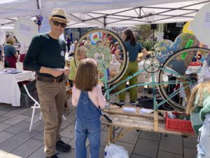Ein Mann mit Hut und Sonnenbrille steht auf einem Jahrmarkt neben einem auf dem Kopf stehenden Fahrrad und spricht mit einem jungen Mädchen mit langen Haaren. Im Hintergrund sind Menschen und Informationsstände unter weißen Zelten zu sehen.