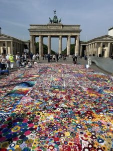 Bunte gehäkelte Blumen bedecken den Boden vor dem Brandenburger Tor in Berlin. An einem sonnigen Tag spazieren die Menschen um die farbenfrohe Installation herum und machen Fotos.