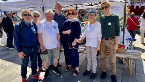 Eine Gruppe von sieben Erwachsenen steht zusammen und lächelt bei einer Veranstaltung im Freien unter weißen Zelten. Einige tragen Sonnenbrillen und Freizeitkleidung, und hinter ihnen sind Informationsstände und bunte Schilder zu sehen.