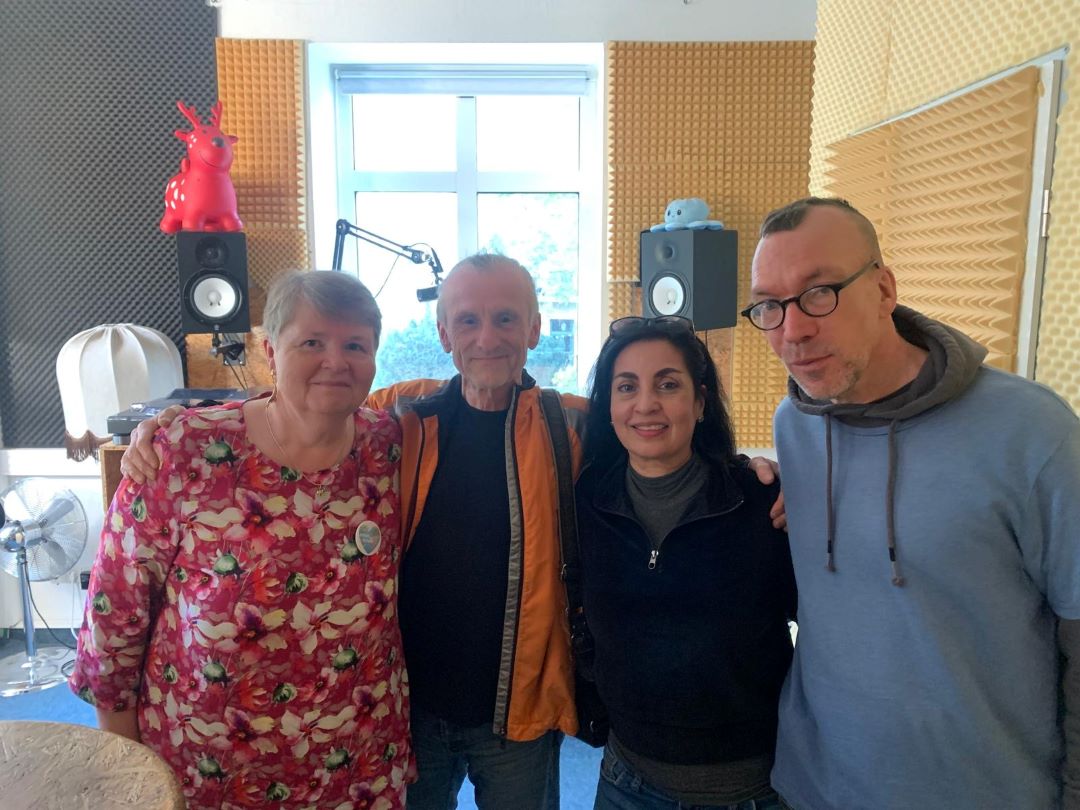 Vier Erwachsene stehen zusammen und lächeln in einem Aufnahmestudio mit schallisolierten Wänden, einem Fenster im Hintergrund und roten und blauen Spielzeugfiguren in Regalen hinter ihnen.
