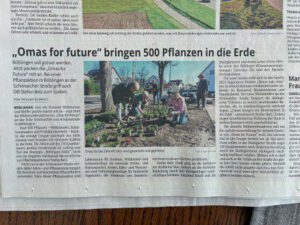 Eine Zeitungsseite mit einer Schlagzeile in deutscher Sprache und einem Farbfoto, das mehrere Personen bei der gemeinsamen Gartenarbeit im Freien zeigt, die in der Nähe eines Gebäudes verschiedene Pflanzen in den Boden pflanzen. Textspalten umgeben das Foto.