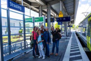 Fünf Personen stehen lächelnd auf einem Bahnsteig unter Schildern mit der Aufschrift Straubing. Neben ihnen hält ein Zug. Die Gruppe wirkt glücklich, eine Person macht ein Selfie. Das Wetter ist klar und sonnig.
