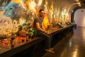 Ein Mann mit Brille und T-Shirt sitzt auf einem schwarzen Tresen in einem Wissenschaftsmuseum, neben dem leuchtende Wandkunst und verschiedene wissenschaftliche Instrumente ausgestellt sind.
