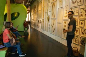 Ein Mann hält Blätter in der Hand und spricht zu einer sitzenden Gruppe in einem Museum, hinter dem eine Wand mit gerahmten botanischen Exemplaren steht. Die Gruppe hört in dem gut beleuchteten, modernen Raum aufmerksam zu.
