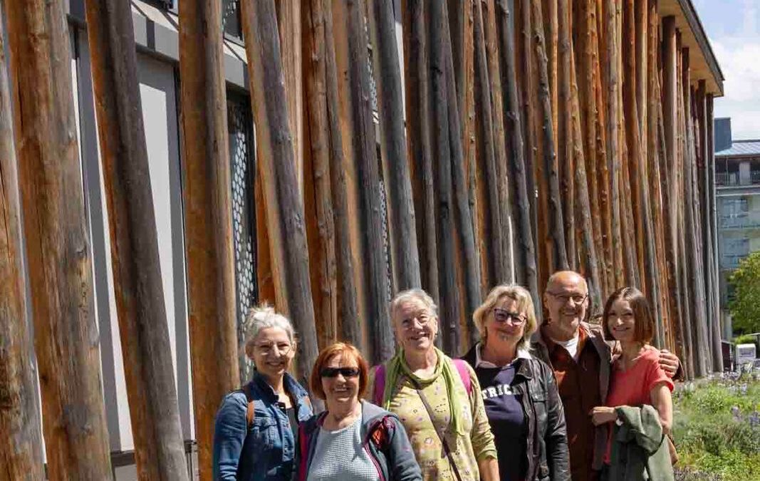 Sechs Erwachsene stehen eng beieinander, lächelnd, vor einem modernen Gebäude mit hohen vertikalen Holzbalken. Sie sind leger gekleidet und genießen einen sonnigen Tag, an dem der Weg neben ihnen von Grün und Blumen gesäumt ist.
