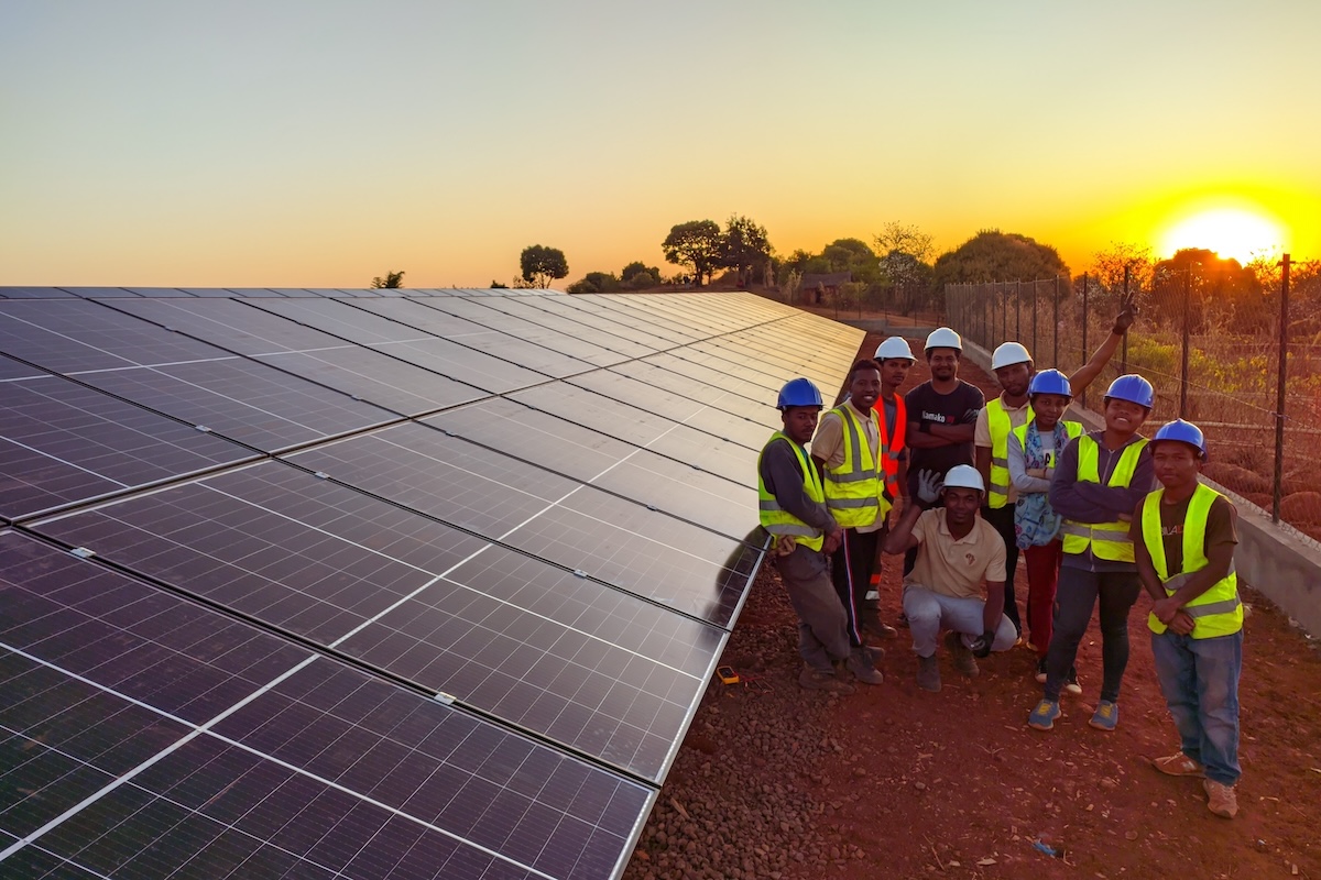 Eine Gruppe von Arbeitern in Sicherheitswesten und Helmen steht bei Sonnenuntergang neben einer großen Solarzellenanlage, die Sonne steht tief am Himmel und die Bäume sind im Hintergrund zu sehen.