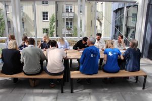 Eine Gruppe von Menschen sitzt an einem langen Holztisch im Freien, einige mit dem Gesicht zueinander, andere mit dem Rücken zur Kamera. Zwei Personen tragen blaue Hemden mit weißen Logos. Im Hintergrund sind Gebäude und Grünflächen zu sehen.