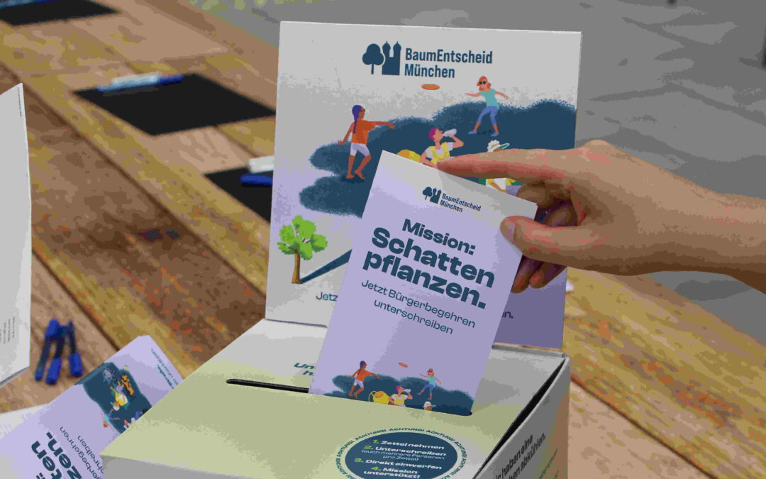 Eine Hand legt ein Flugblatt in eine Box mit der Aufschrift "Mission: Schatten pflanzen" und "baumentscheid münchen" und wirbt damit für die Münchner Baumpflanzaktion. Die Szene spielt sich im Freien auf einem Holztisch ab.